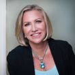 Woman with blonde hair wearing a black blazer and necklace smiling in front of a white wall.