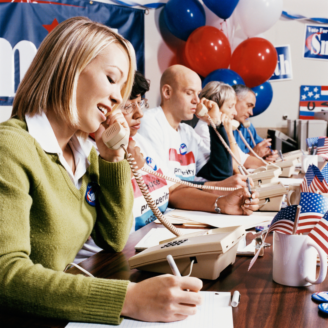 People making calls in a campaign office. Balloons, flags, and signs are visible.