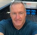 Man in blue shirt smiles, looking at the camera. He is in the back of a truck, toolbox behind him.