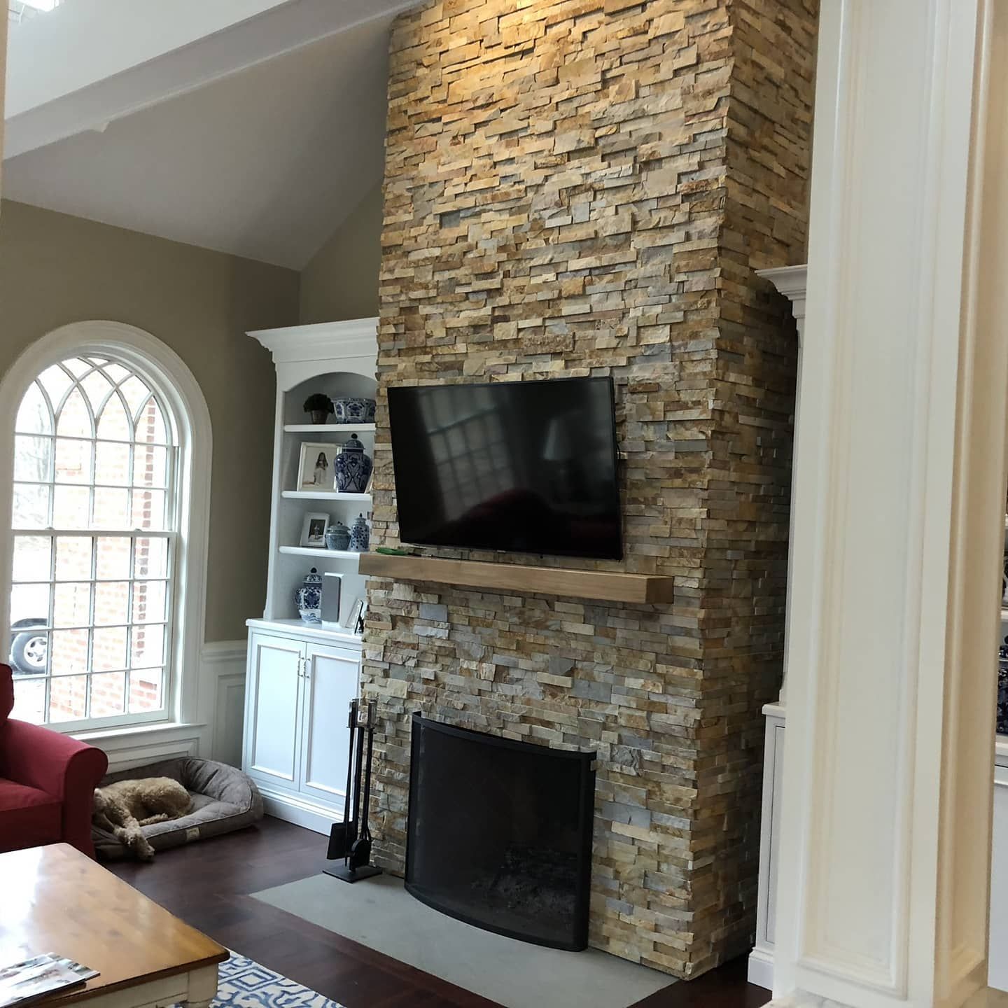 Living room with stone fireplace, TV, built-in shelving, arched window, and dog bed.