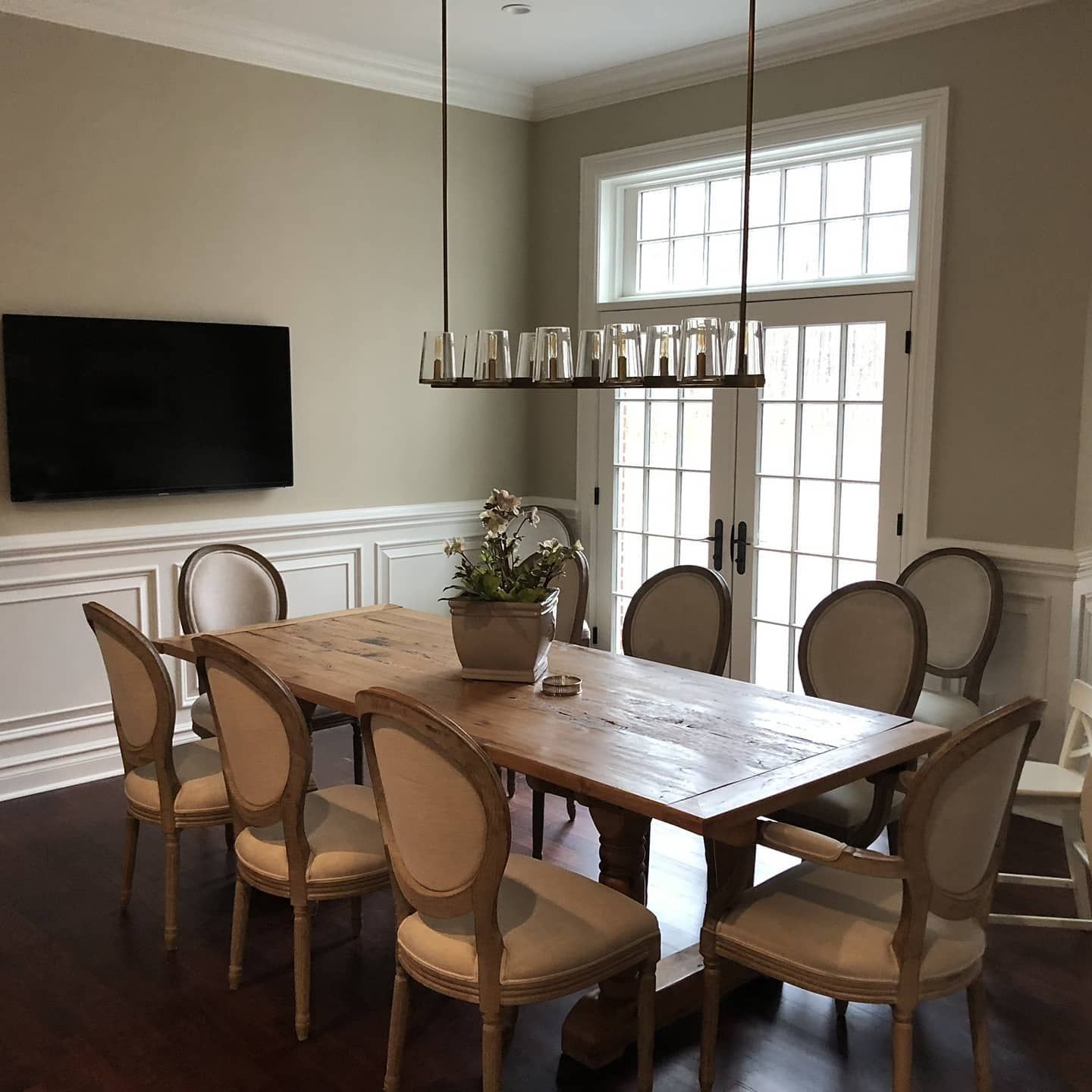 Dining room with wooden table, eight upholstered chairs, and a chandelier. A TV is mounted on the wall.
