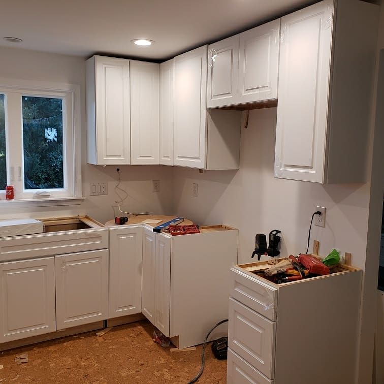 White kitchen cabinets being installed in a room with a window, tools, and exposed flooring.
