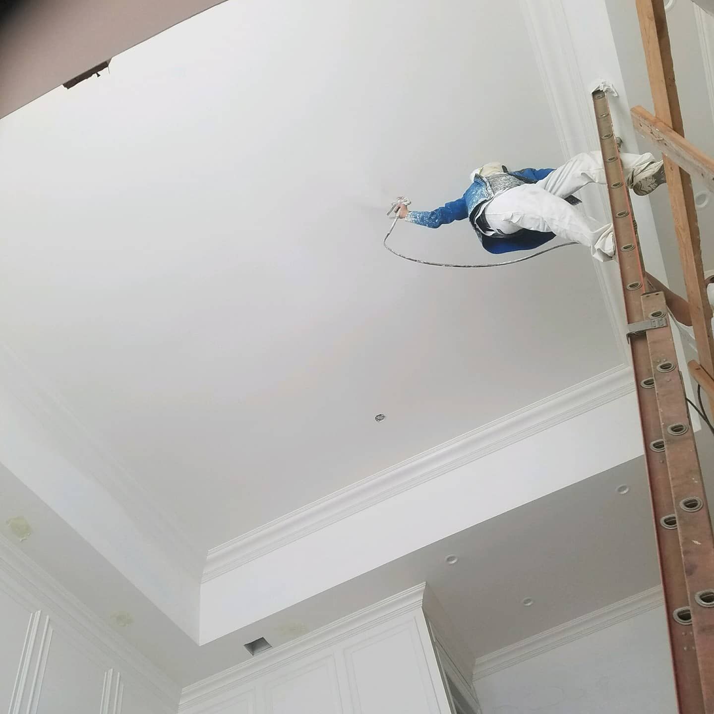 Person painting a white ceiling with a spray gun, standing on a ladder.