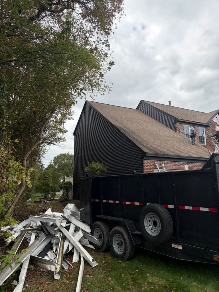 Black dumpster trailer next to a house with brown roof and white debris pile, under an overcast sky.