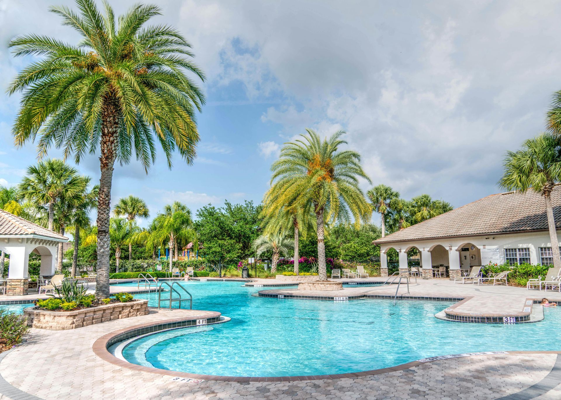 Swimming pool with palm trees, blue water, and a building with a thatched roof, sunny day.