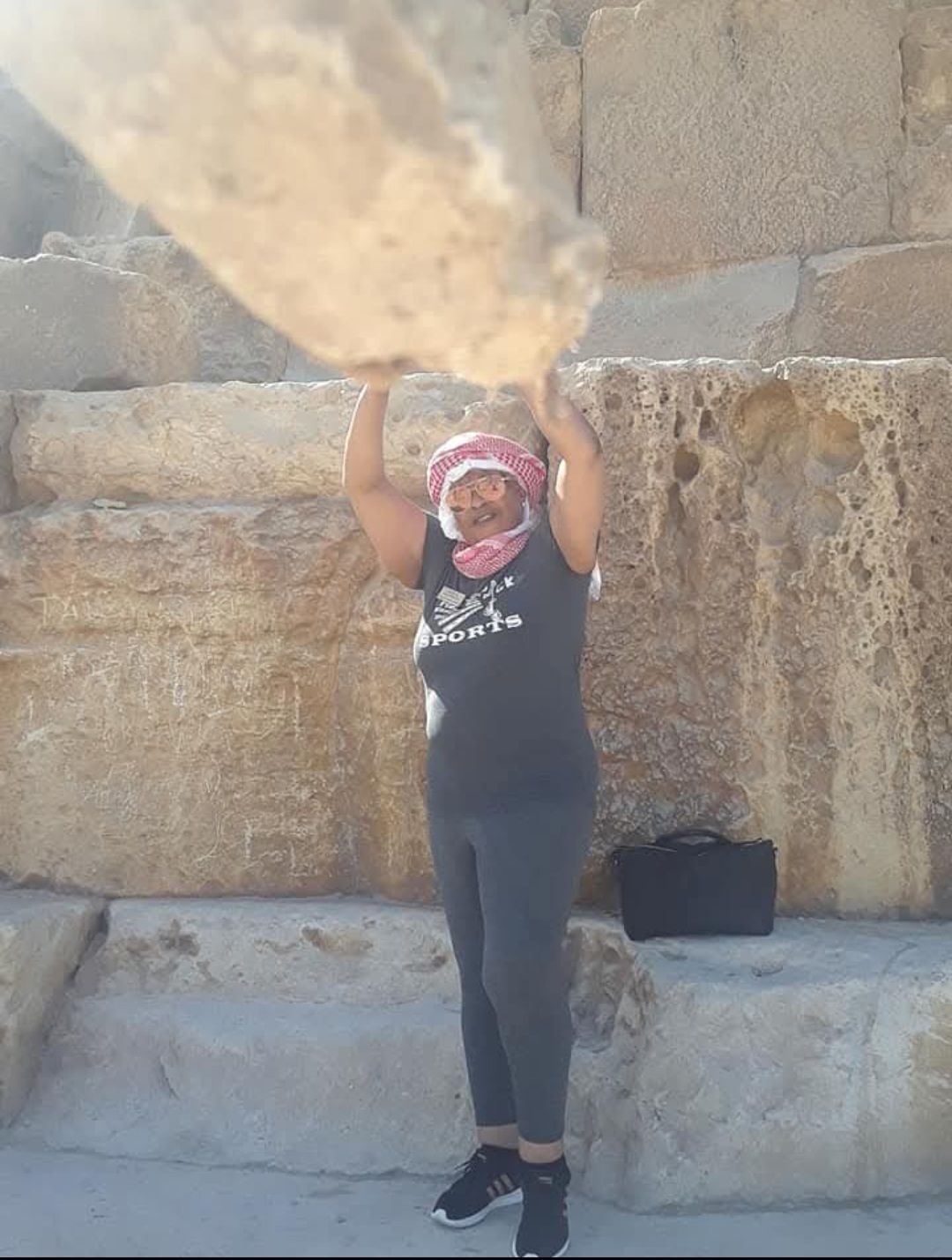A woman is standing in front of a stone wall holding a large rock.