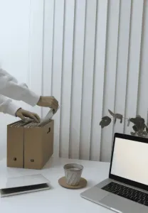 Pessoa arquivando papéis em uma caixa de papelão sobre uma mesa branca com um laptop, persianas e uma xícara de café.