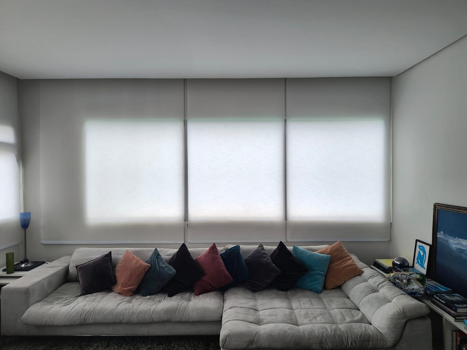 Um sofá secional cinza com almofadas coloridas em frente a três persianas que filtram a luz.