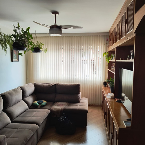 Sala de estar aconchegante com sofá marrom, centro de entretenimento de madeira e plantas penduradas perto de uma grande janela com persianas verticais.
