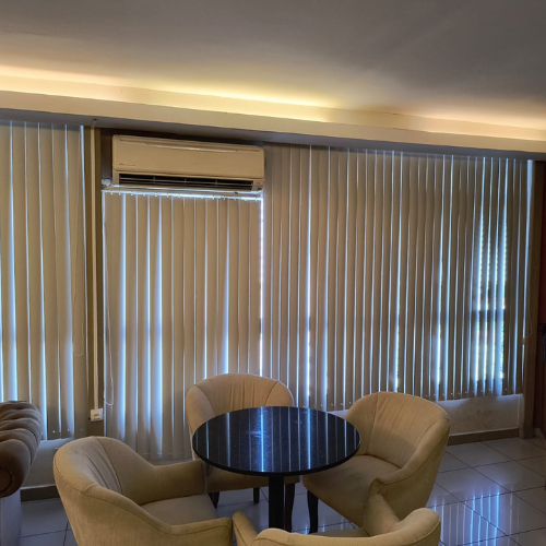 Uma sala com uma pequena mesa redonda e quatro poltronas bege perto de uma janela com persianas verticais.