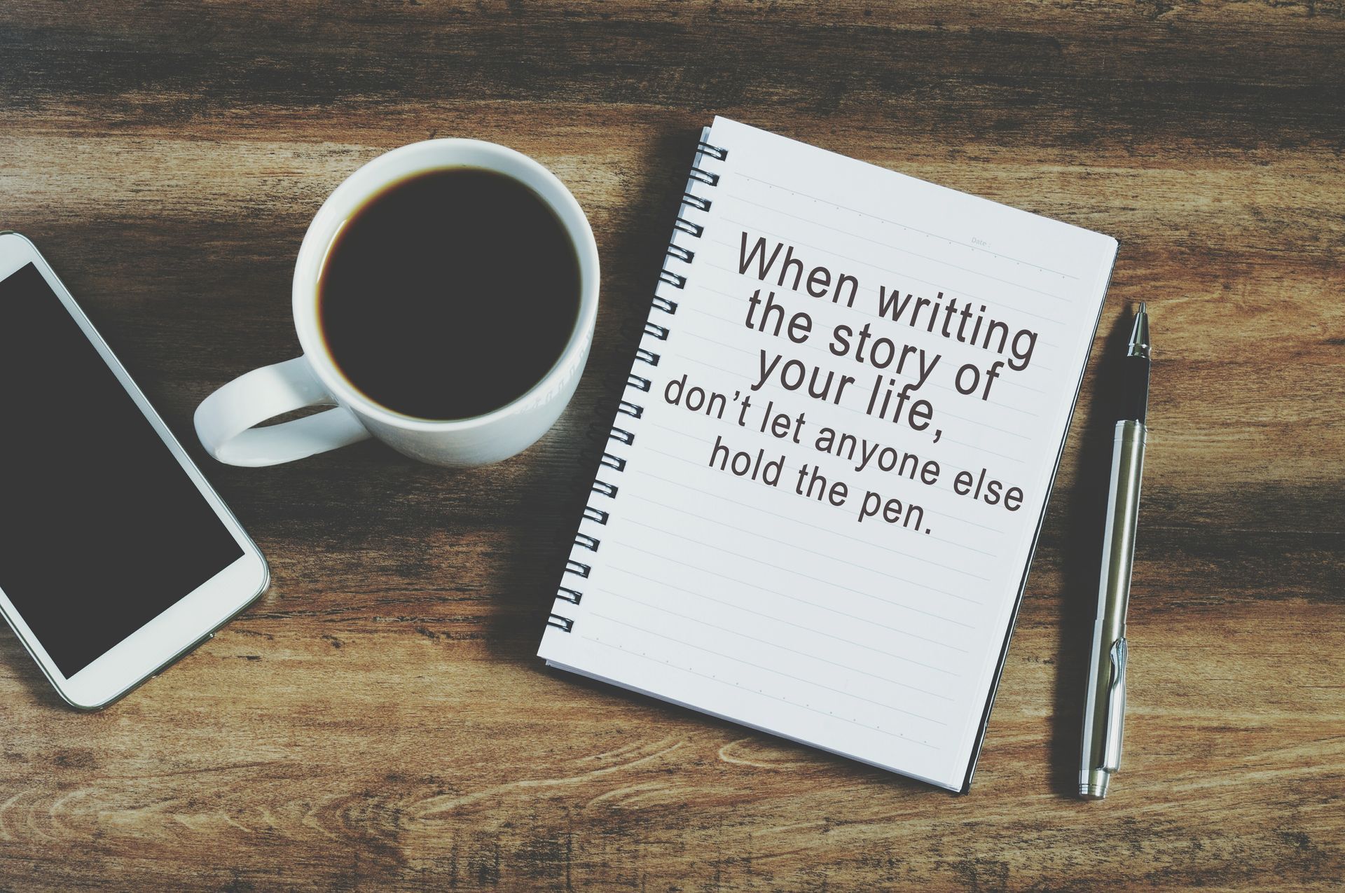 Notebook with quote, coffee, pen, and phone on wood.