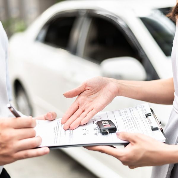 Hands exchanging car keys and paperwork beside a white car at a dealership