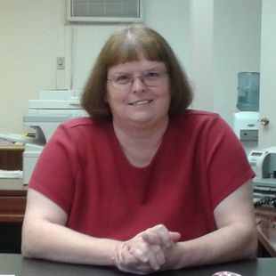 Person in a red shirt smiling and leaning on a desk in an office setting