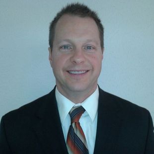 Smiling person in a black suit, white shirt, and striped tie against a plain light background