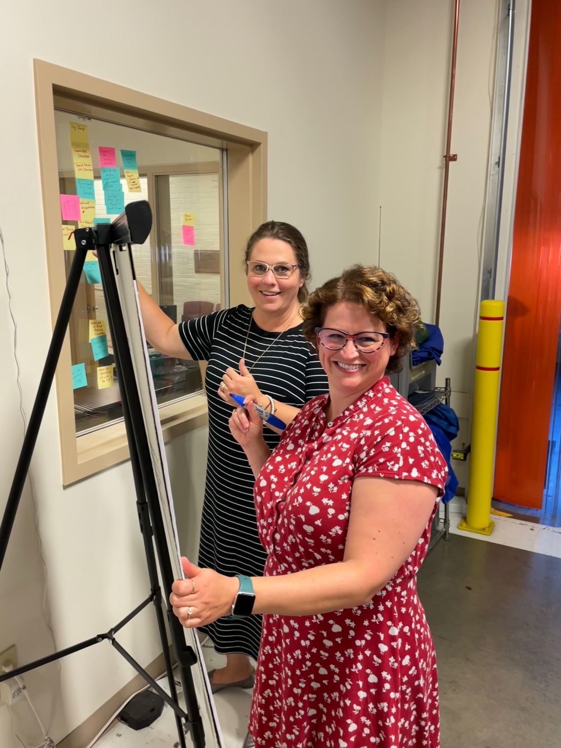 Two women are standing next to each other in front of a window with sticky notes on it.