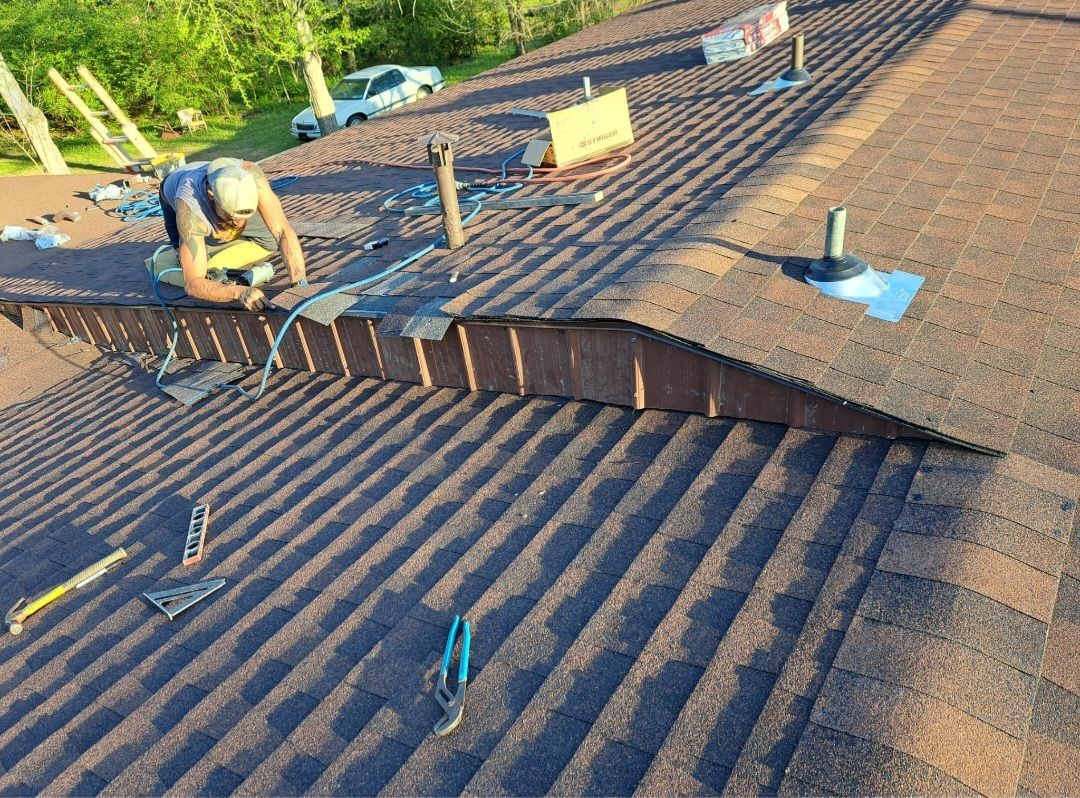 Workers repairing a brown shingled roof with ladders and tools on a sunny day