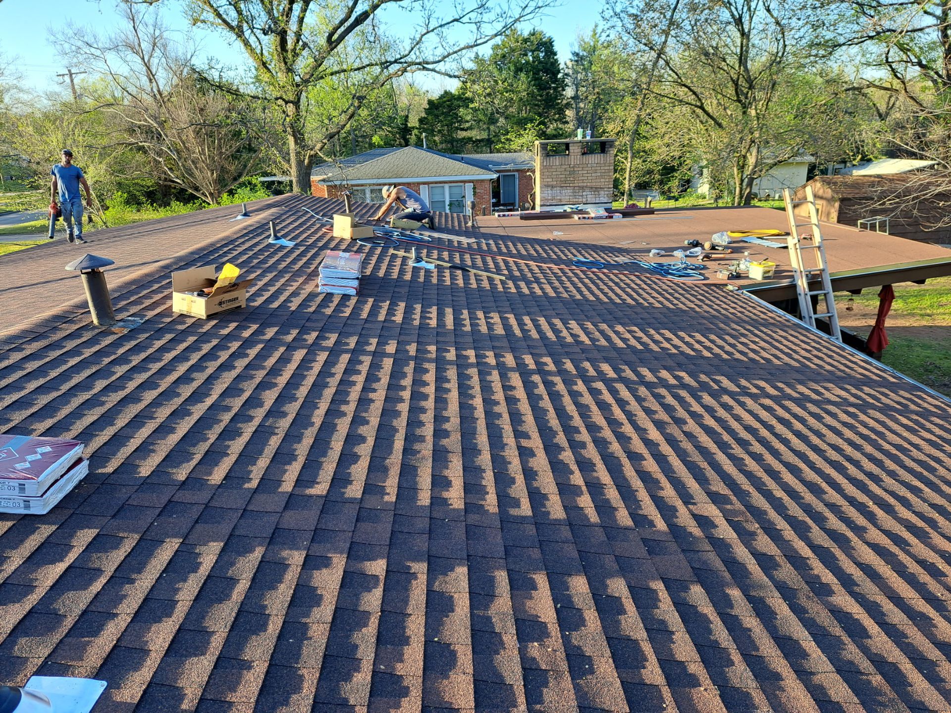 Worker repairing a shingled roof beside a brick chimney under a blue sky