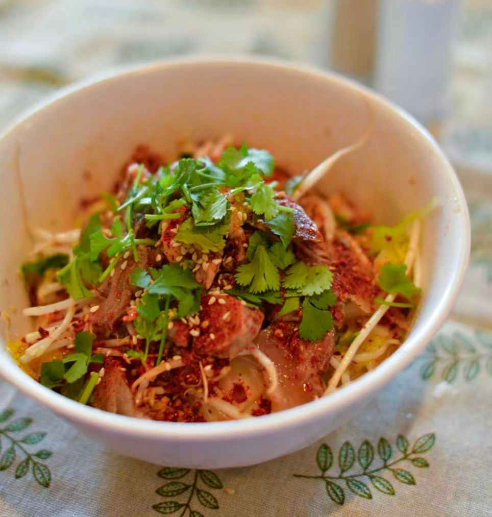 White bowl of noodles topped with herbs and spices on a patterned tablecloth