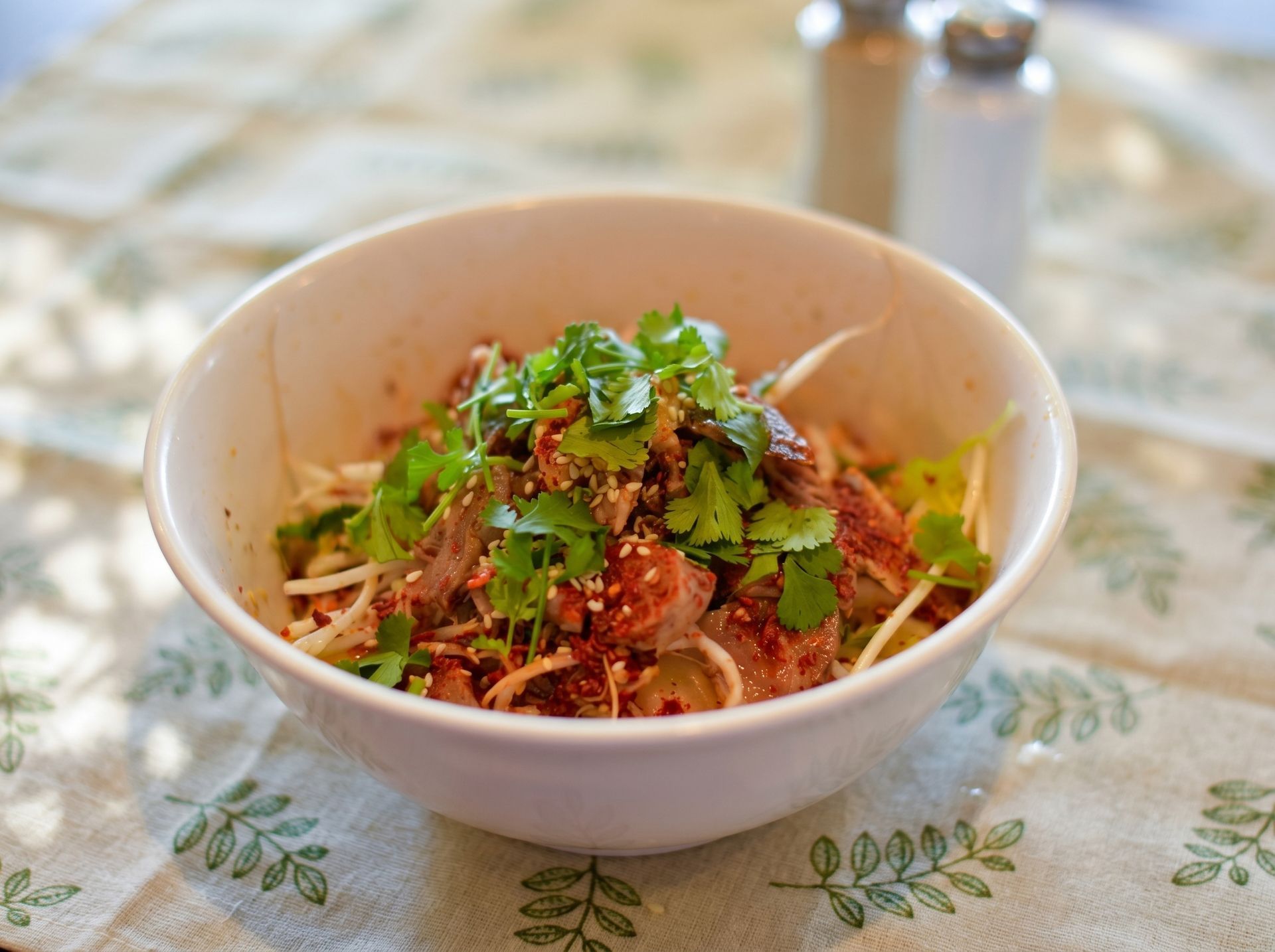 White bowl of noodles topped with herbs and spices on a patterned tablecloth