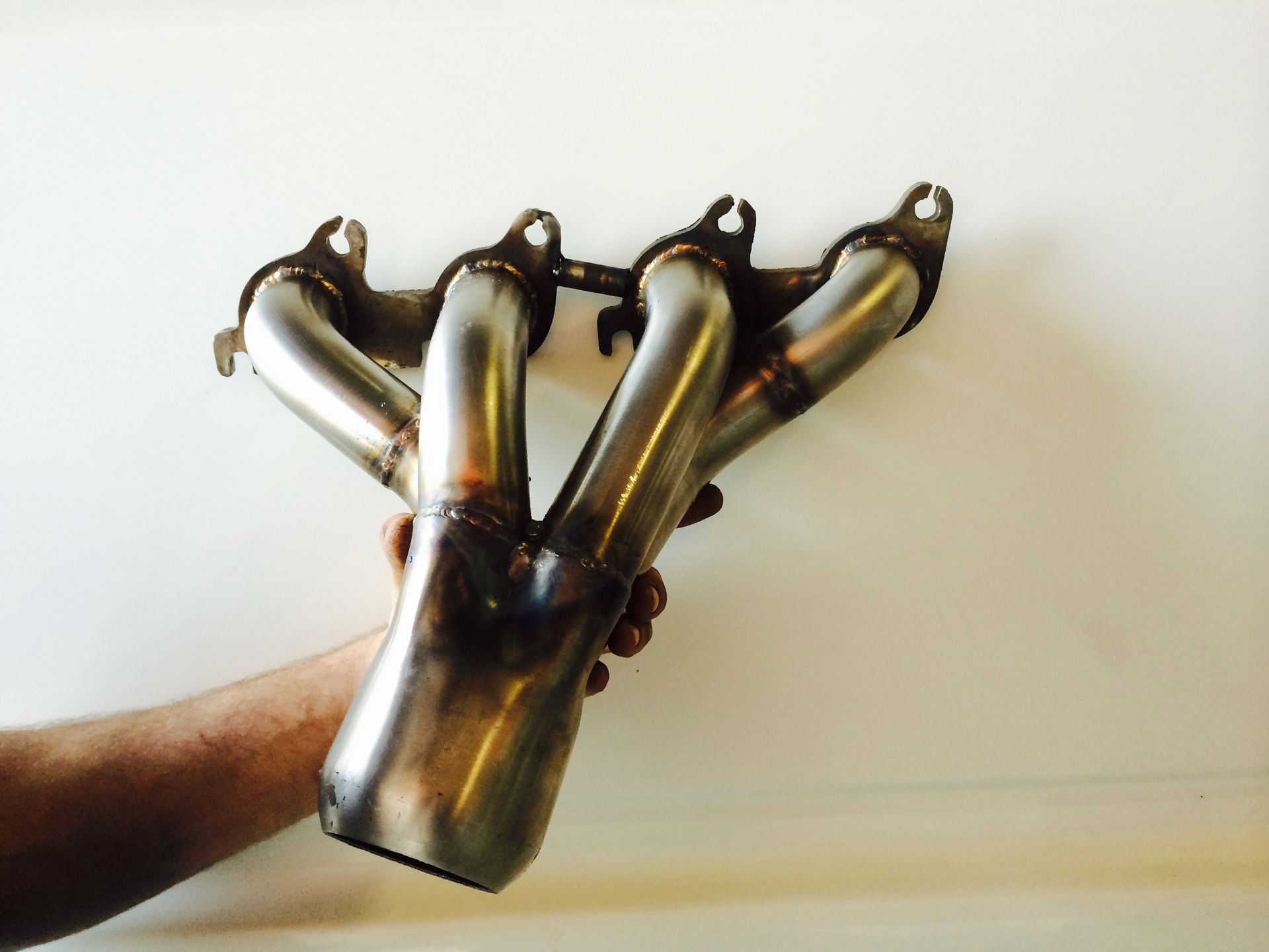 Hand holding a shiny metal exhaust header. It has four tubes converging into one, with mounting flanges.