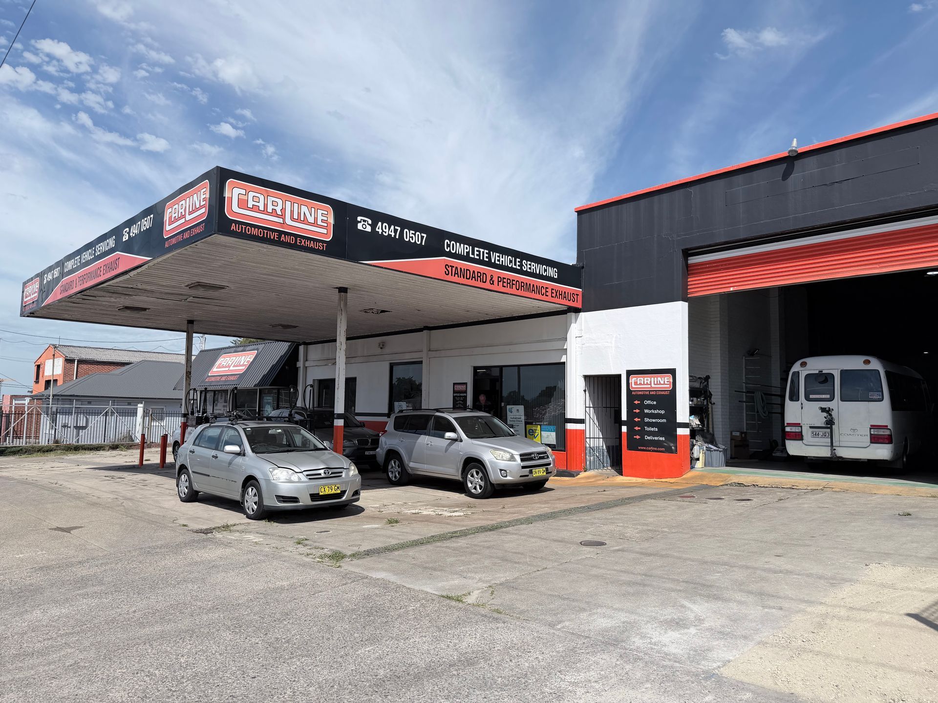 Gas station with red and black signage, silver cars parked under a canopy. White van in a bay.
