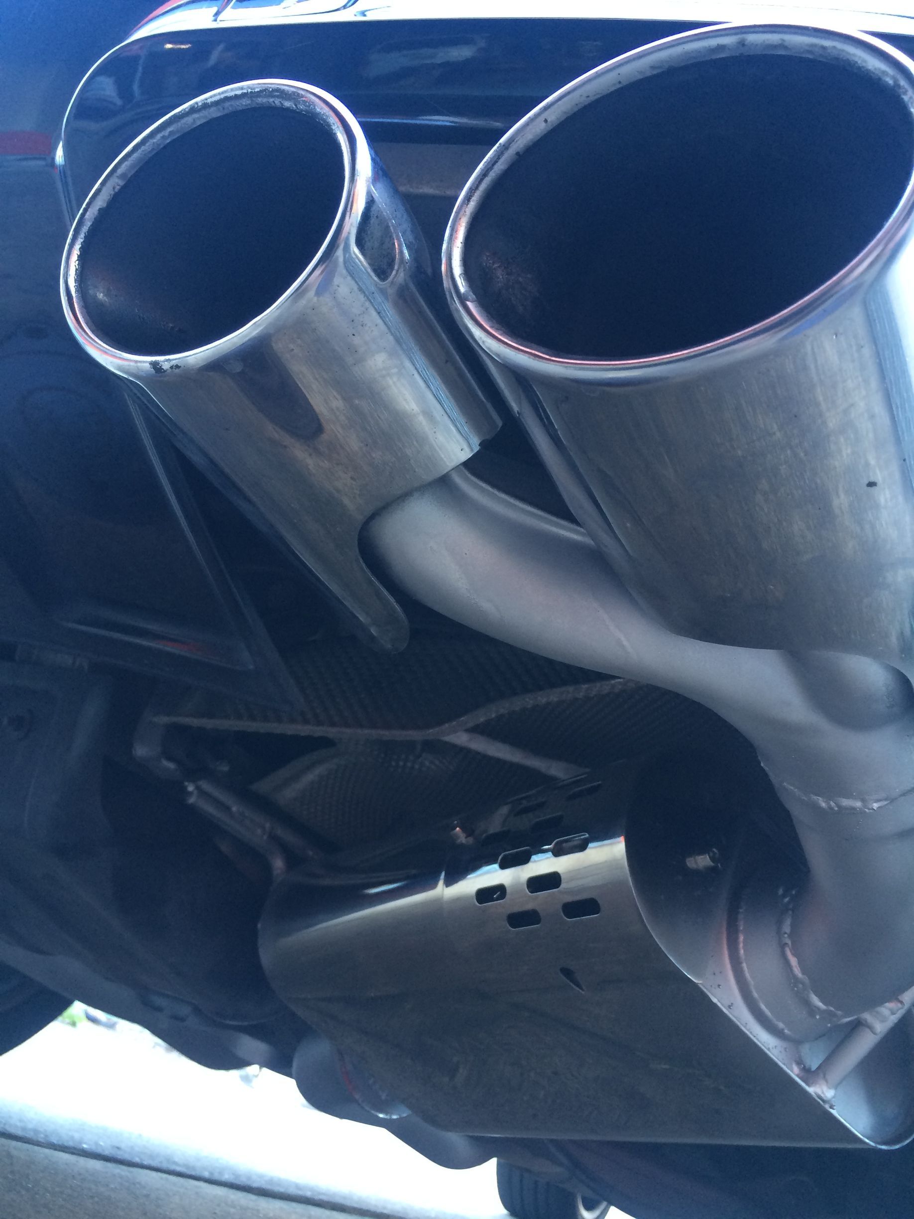 Chrome exhaust pipes under a car, viewed from below. Two large, round tips and a rectangular muffler.