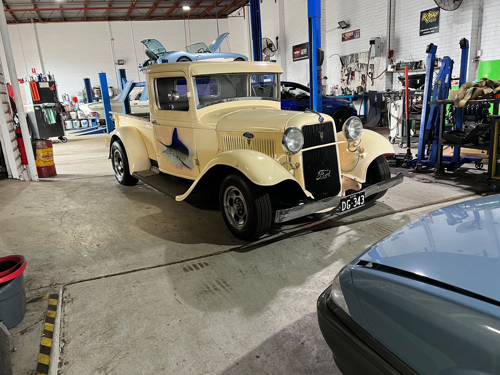 Classic cream-colored pickup truck inside a garage, with a shark graphic, and another car visible in the foreground.