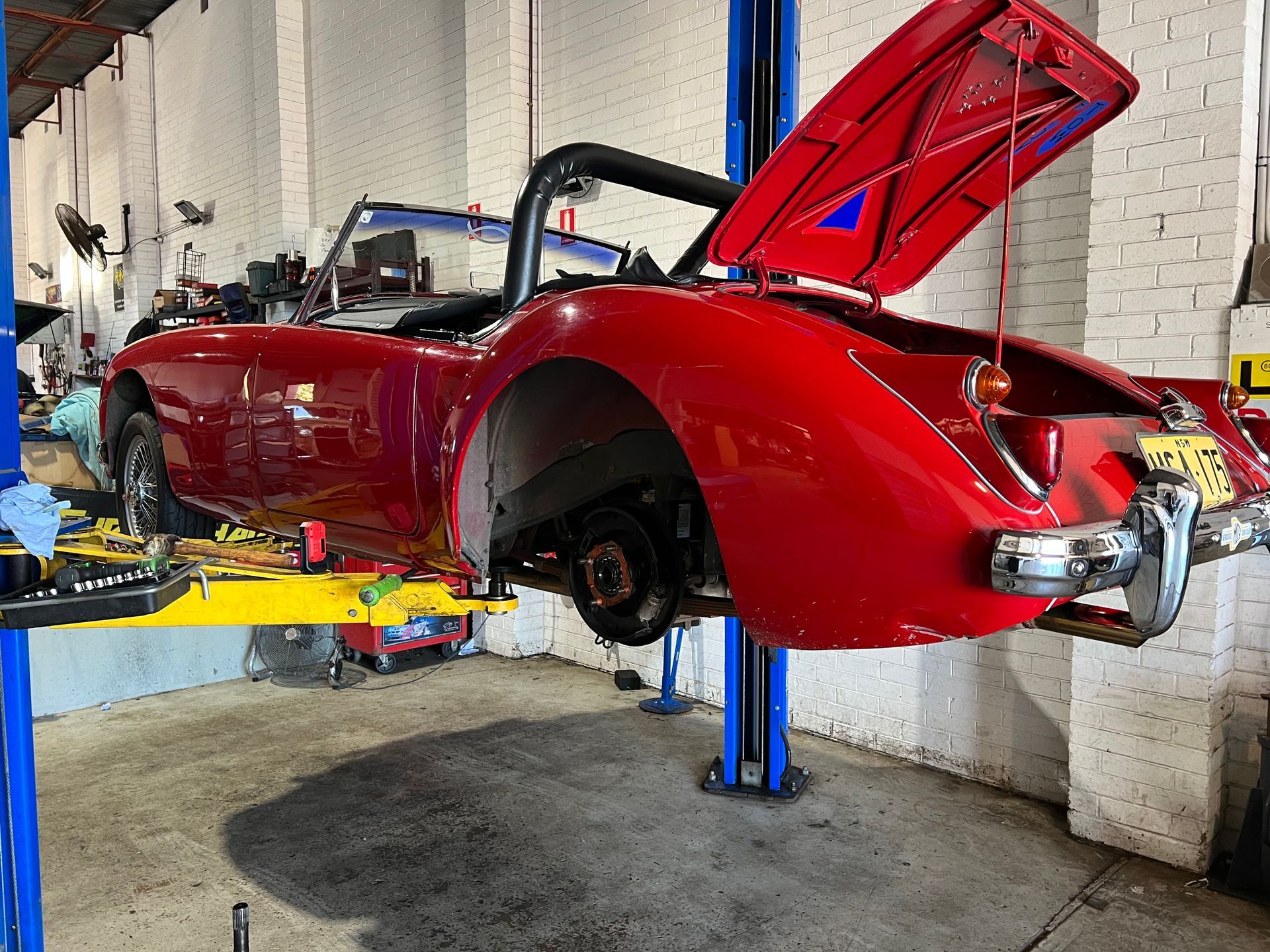 Red classic car on a lift, rear open. Car is in a garage with tools visible.