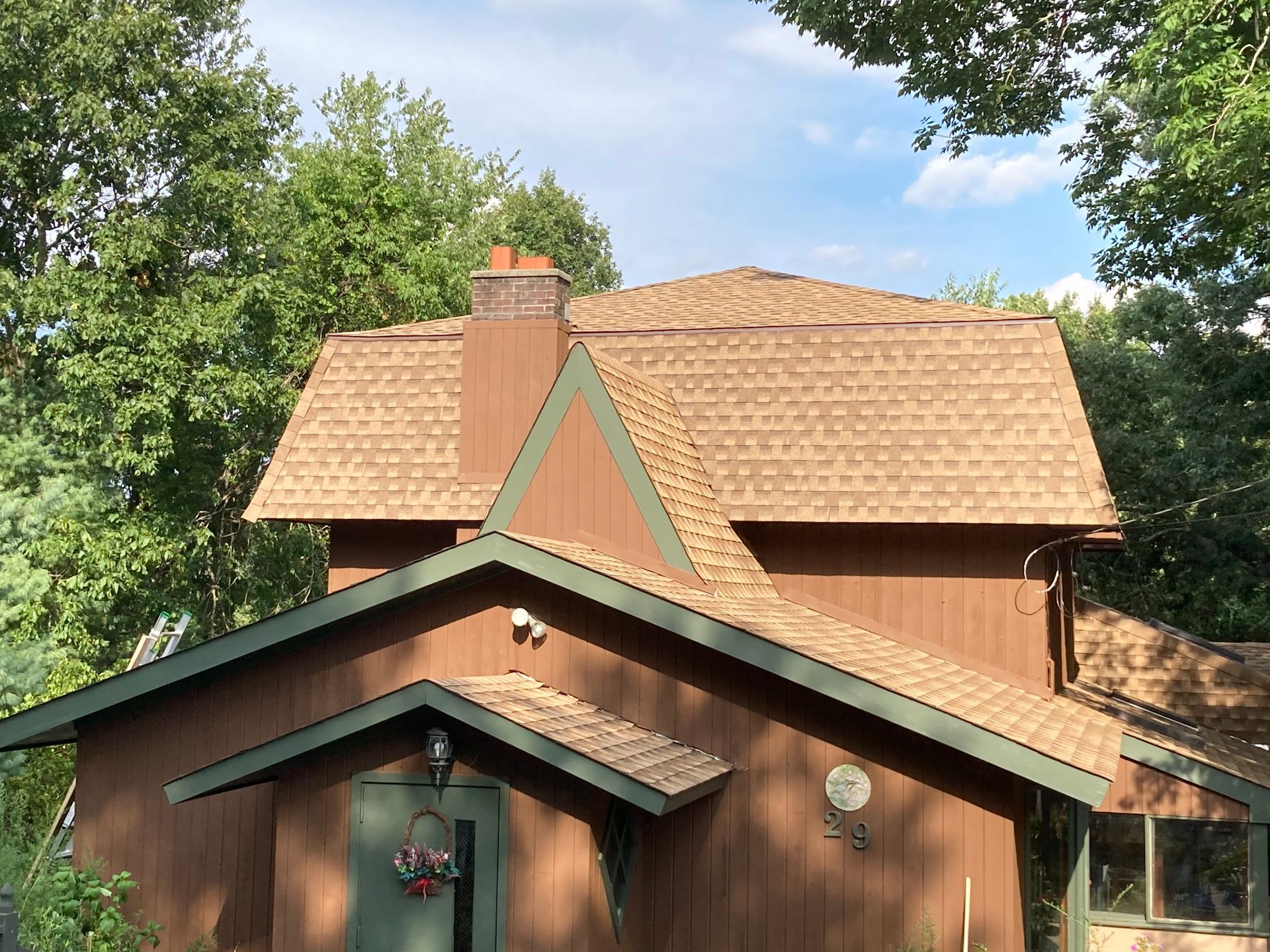 A two-story brown house with a shingled roof, a chimney, and a small front entrance porch, set among green trees.