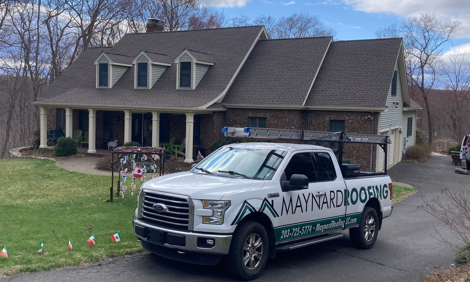 A white Maynard Roofing pickup truck parked in front of a house with a shingled roof and a front porch.