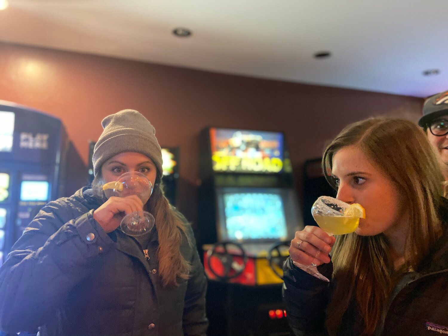 Two women drinking from glasses in front of an arcade machine