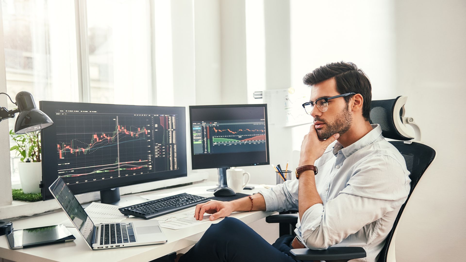 Pessoa analisando gráficos de ações em dois monitores em uma mesa em um escritório bem iluminado.
