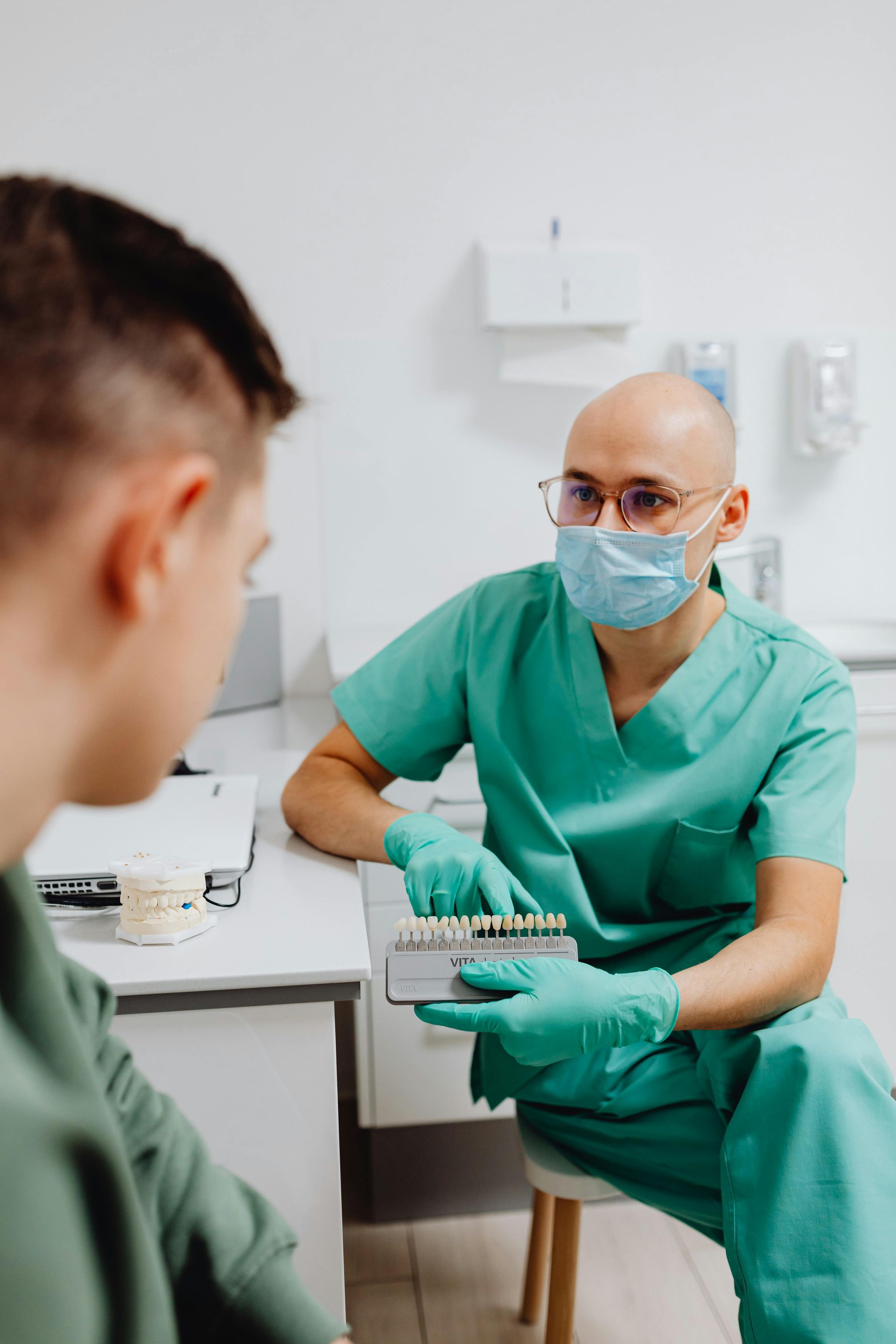 Dentista de uniforme cirúrgico mostra tabela de cores para paciente em consultório odontológico.