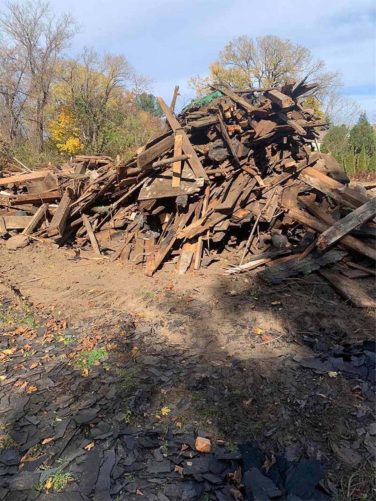 Pile of weathered wood debris on muddy ground, trees in the background under a blue sky.