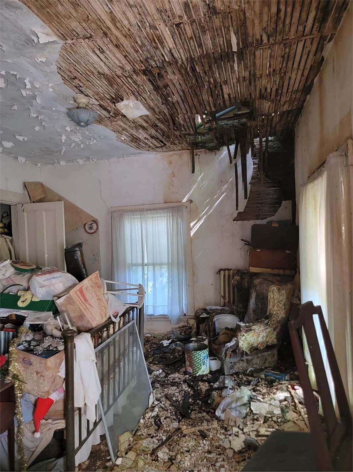 Damaged, cluttered room with collapsing ceiling, debris, and window with curtains.