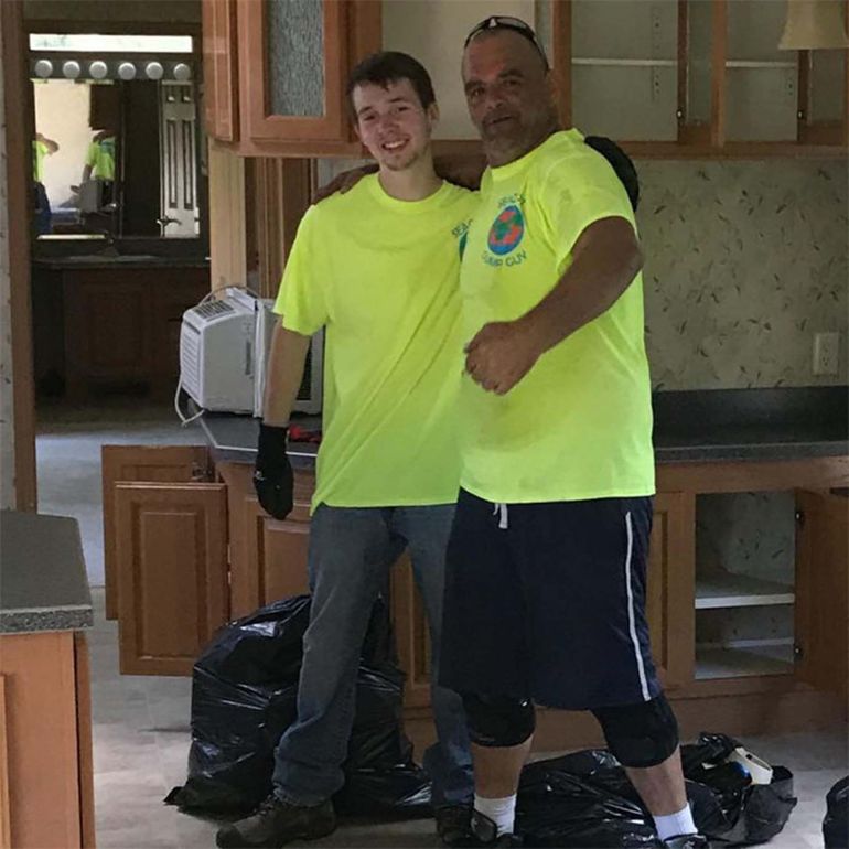 Two men in neon shirts stand in a room, arm in arm. Trash bags and cabinets are visible.