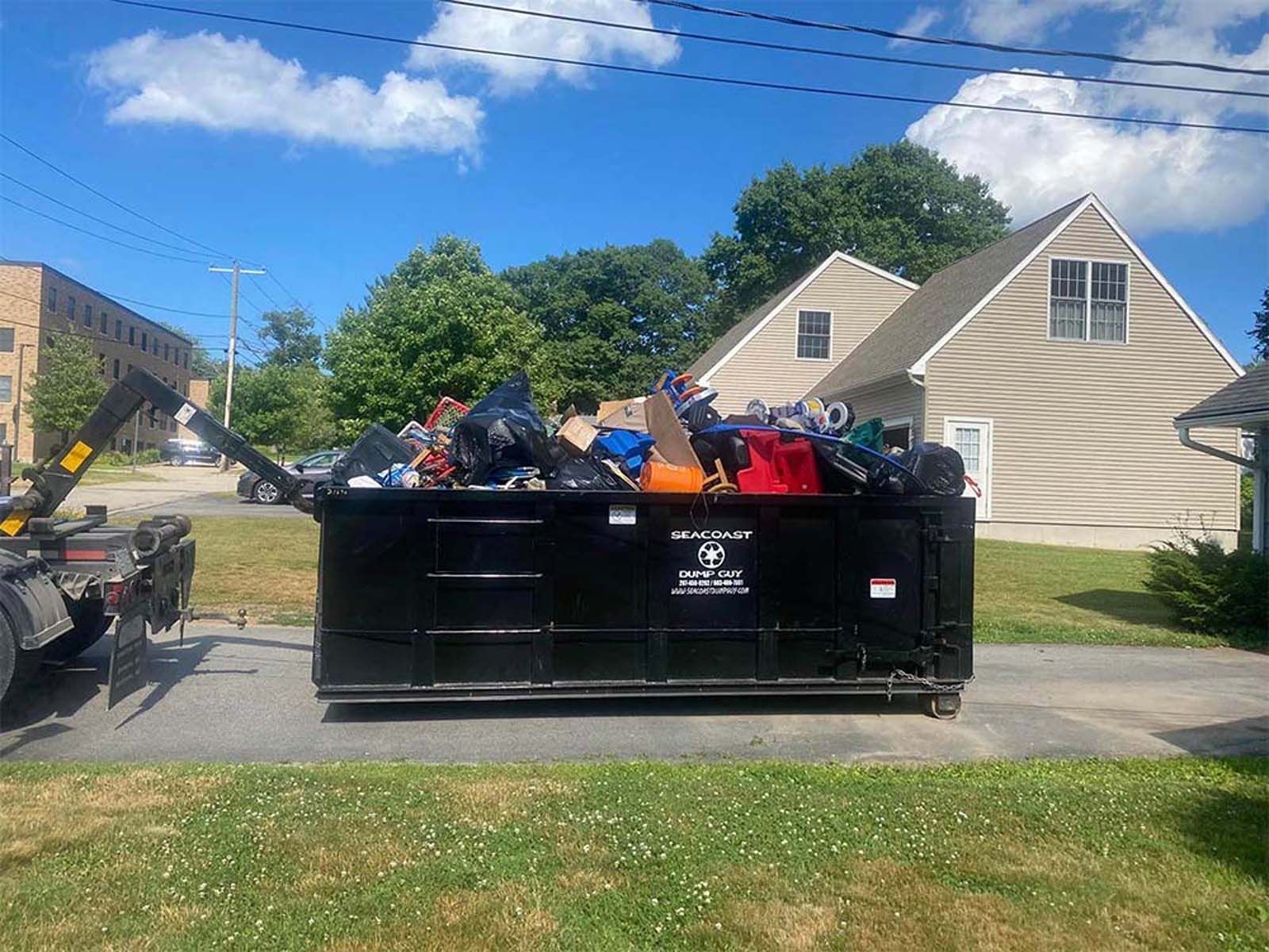 Black dumpster overflowing with trash on a sunny day in front of a house.