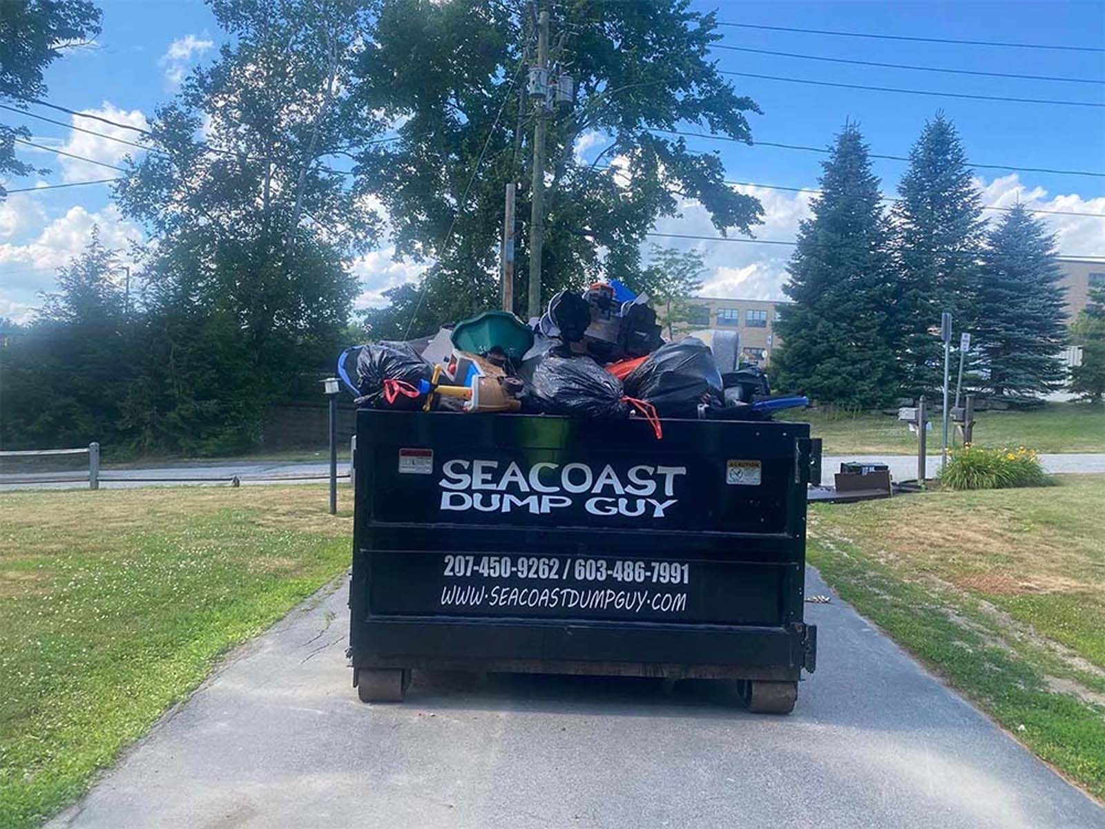 Black dumpster filled with trash bags on a paved driveway; Seacoast Dump Guy logo visible.