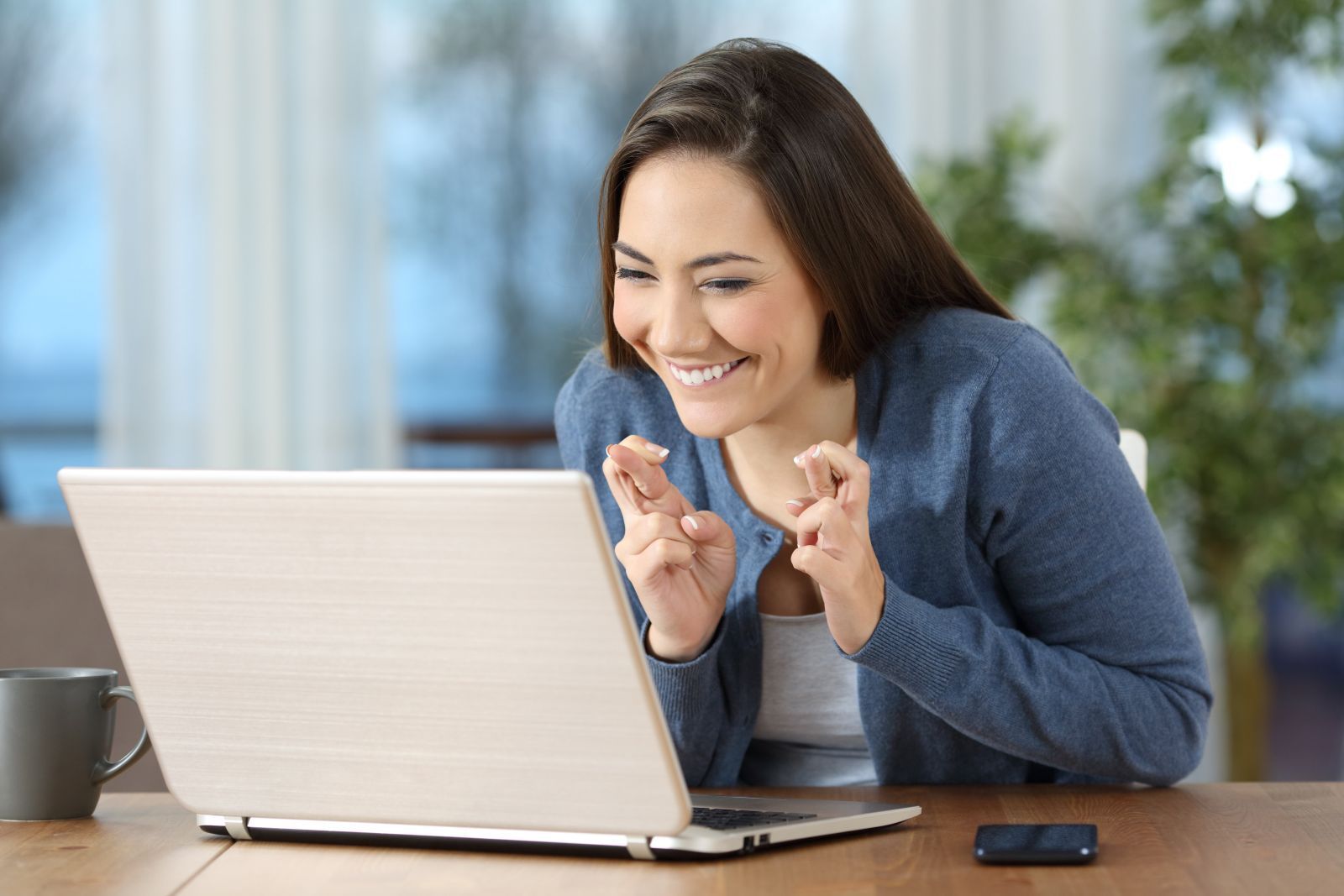 A woman is sitting at a table using a laptop computer and has her fingers crossed while she's smiling