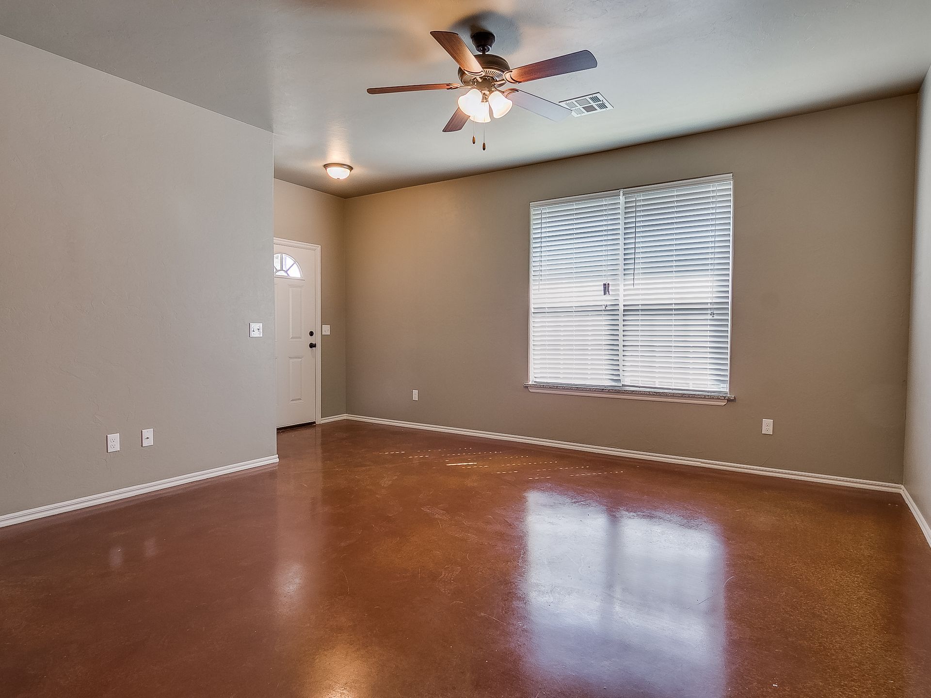 Empty living room with brown floor and taupe walls, ceiling fan, window with blinds.