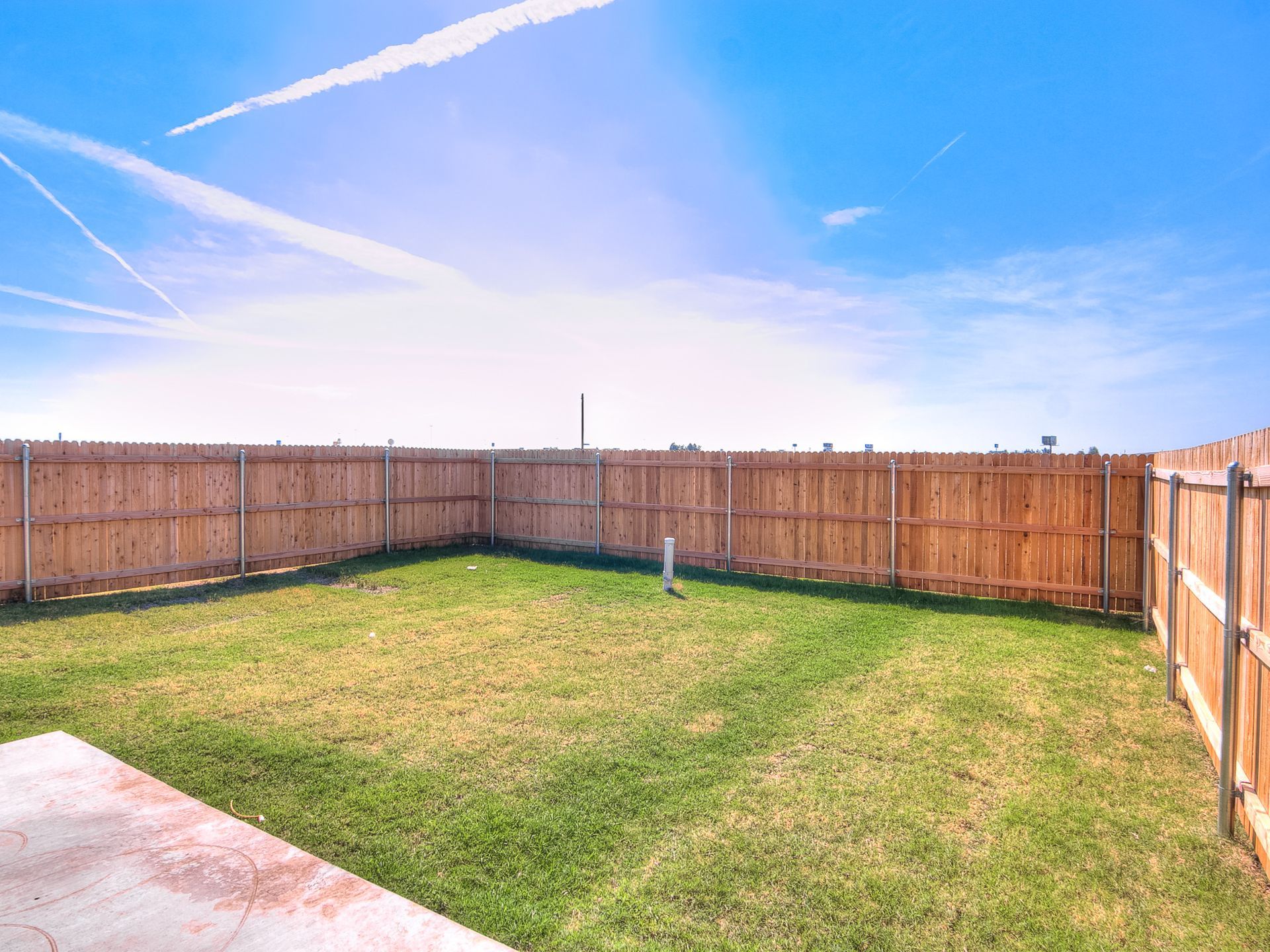Grassy backyard enclosed by a wooden fence under a bright blue sky with contrails.