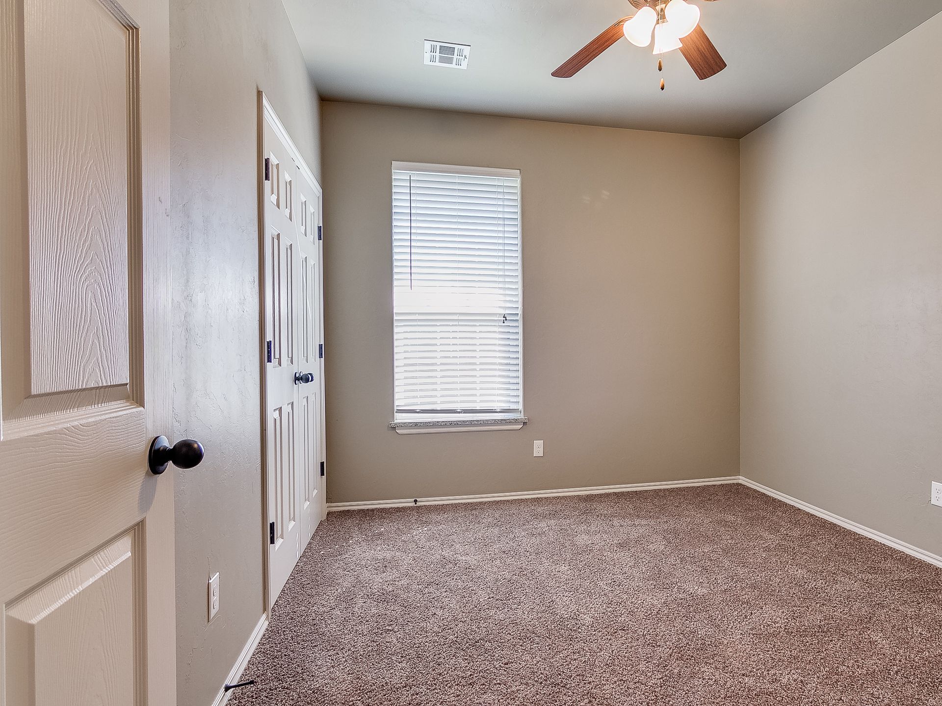 Empty bedroom with brown carpet, tan walls, window with blinds, and a closed door.