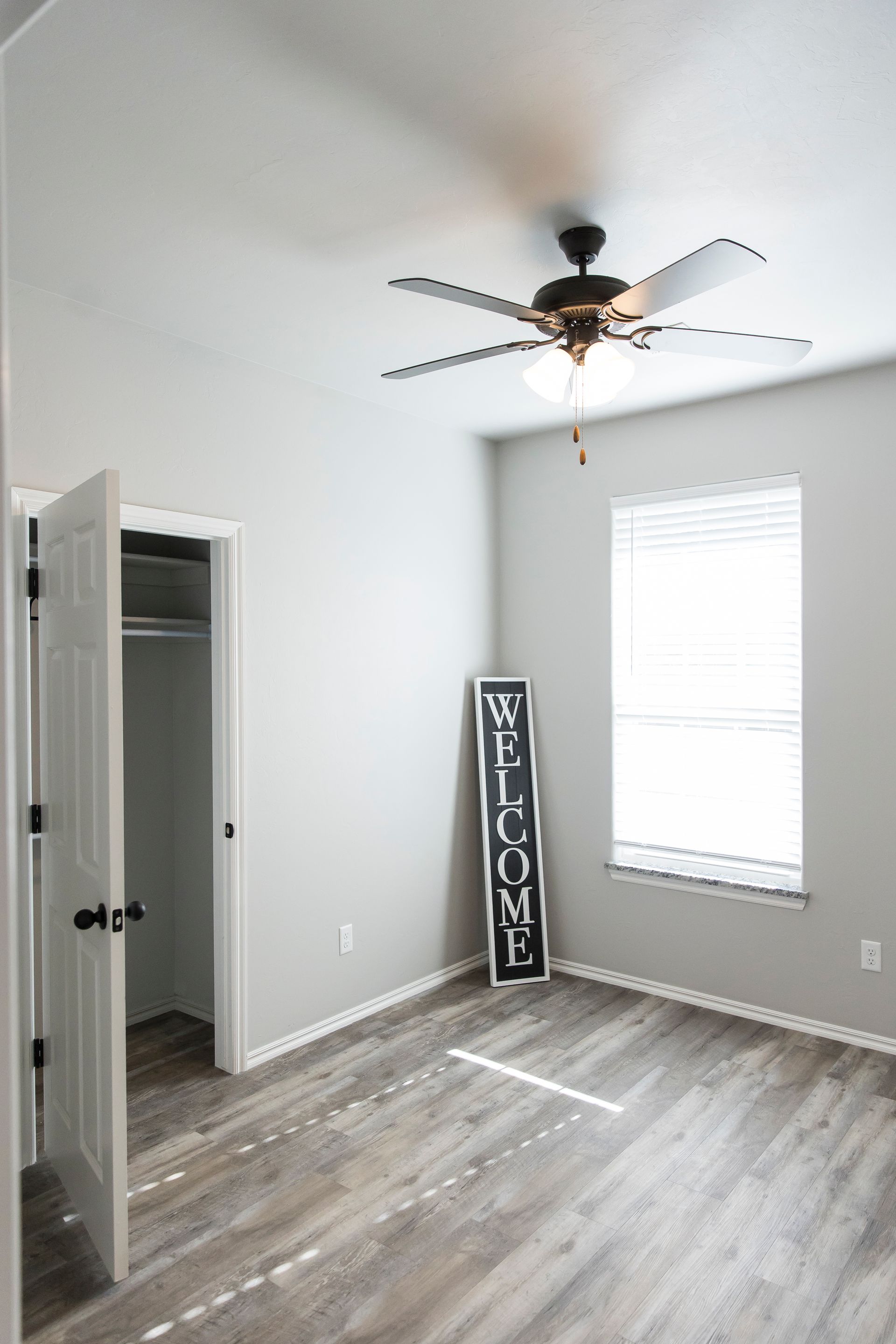 Bedroom with gray walls, wood-look floors, a closet, window, and a Welcome sign. A ceiling fan is installed.