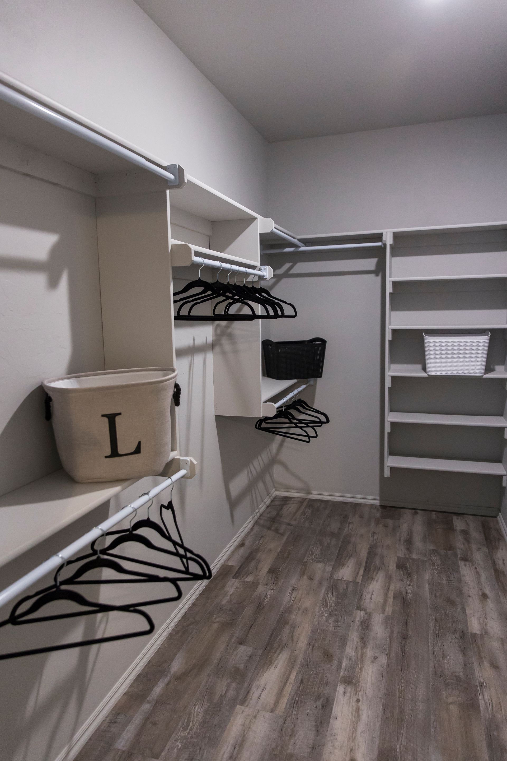 Walk-in closet with white shelves, hanging rods, and gray wood-look flooring. A basket with