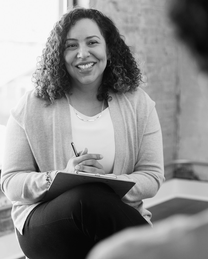 Woman with curly hair smiles, holding a clipboard, facing a person seated out of view.
