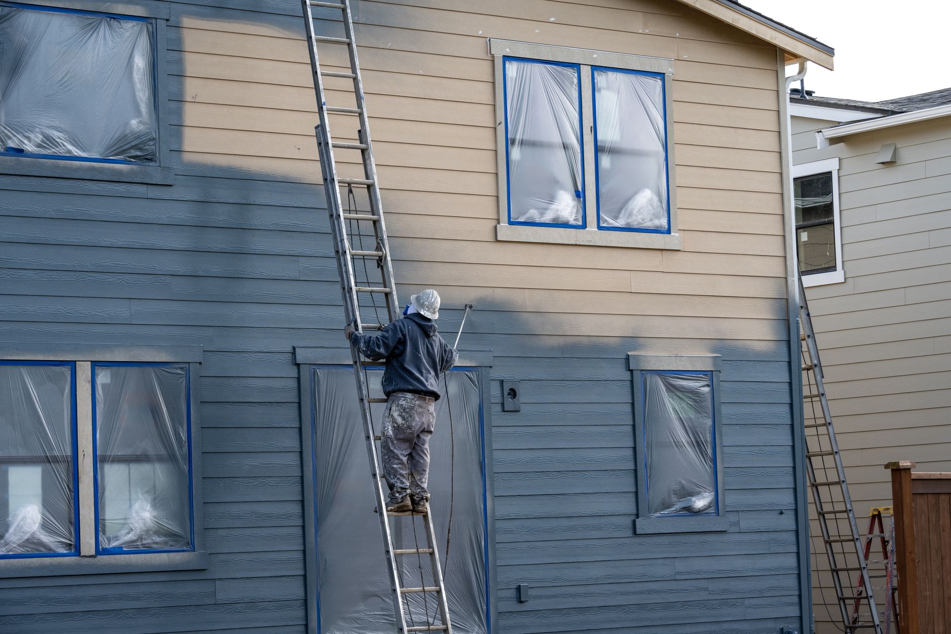Person painting a house, blue paint on half the wall, windows and door covered, ladder.