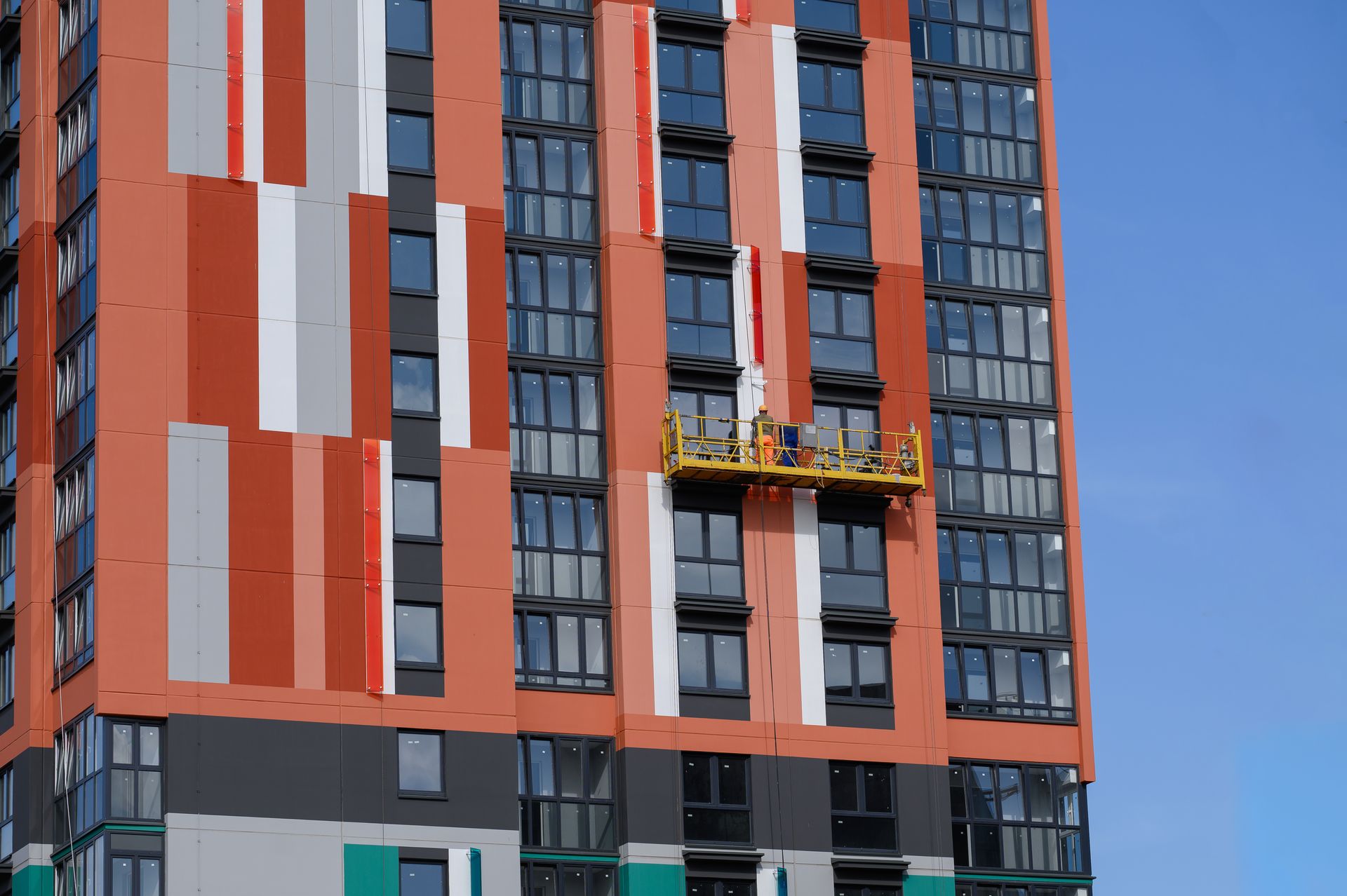 High-rise building with orange, grey, and white panels; workers on a suspended platform; blue sky.