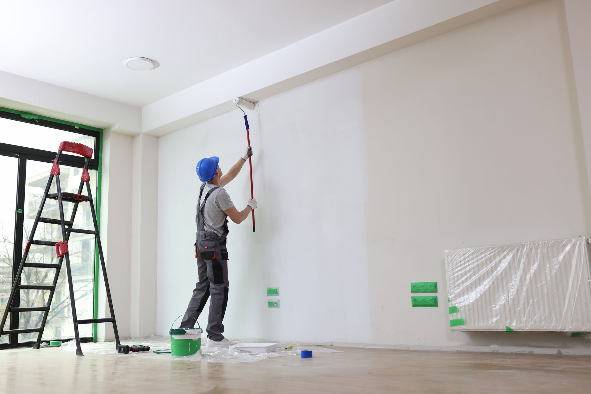 Person in overalls and blue hardhat painting a white wall with a roller. Interior room with ladder, paint can.