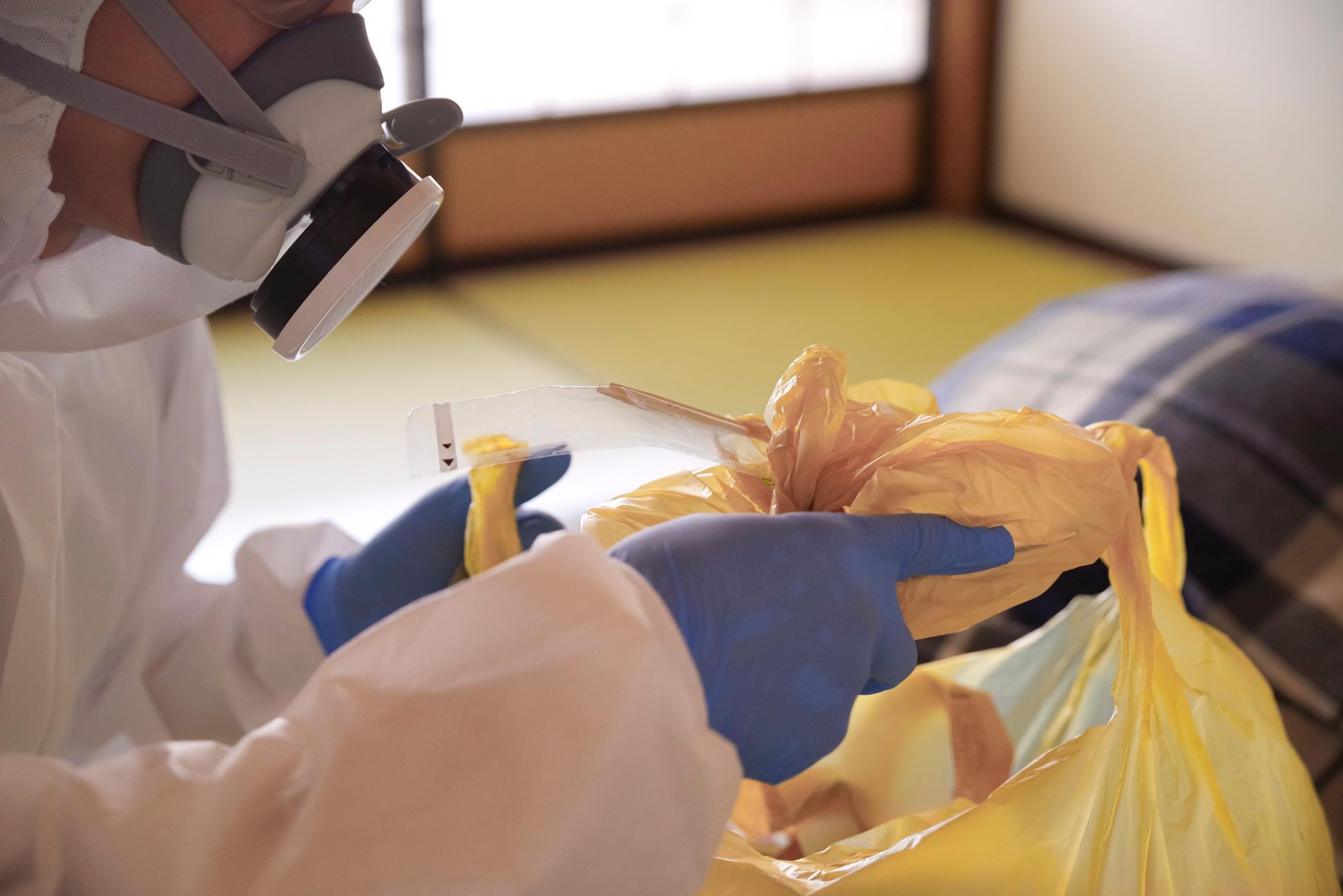 Person in gloves and mask handling a yellow trash bag in a room.