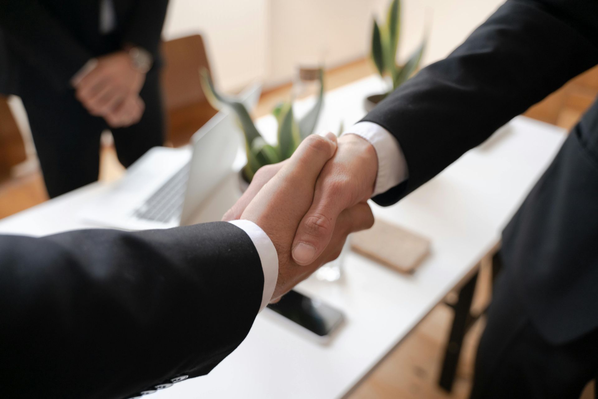 Two people in suits shaking hands at a white table, with a laptop and plants in the background.